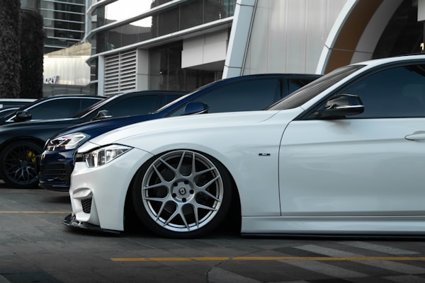 Several luxury cars are parked in a row in an urban setting with modern architecture in the background. The focus is on a sleek white car with custom wheels and low suspension.
