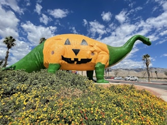 A large dinosaur sculpture painted like a jack-o'-lantern stands prominently in a landscaped area filled with yellow and orange flowers. The dinosaur is predominantly green with an orange pumpkin-like body, black eyes, nose, and a carved grin. Palm trees are visible in the background under a partly cloudy sky, with mountains further in the distance. Several cars are parked along a nearby road.