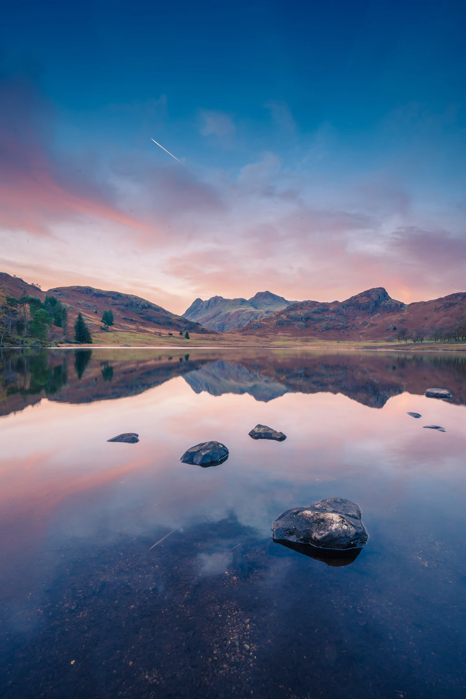 Serene lake surrounded by mountains with clear blue sky and distant airplane smoke trail