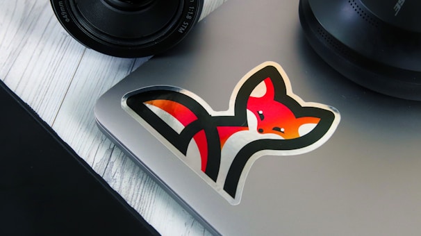 A laptop with a colorful fox sticker on it, positioned on a white wooden surface. Nearby are parts of a camera lens and a circular speaker.