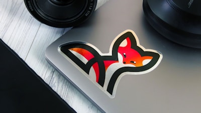 A laptop with a colorful fox sticker on it, positioned on a white wooden surface. Nearby are parts of a camera lens and a circular speaker.