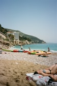 A sunny beach with several kayaks lined up on the shore, people enjoying the water and relaxing on the sand. Hills and buildings provide a scenic backdrop, and the sea is a vibrant blue.