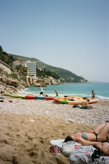 A sunny beach with several kayaks lined up on the shore, people enjoying the water and relaxing on the sand. Hills and buildings provide a scenic backdrop, and the sea is a vibrant blue.