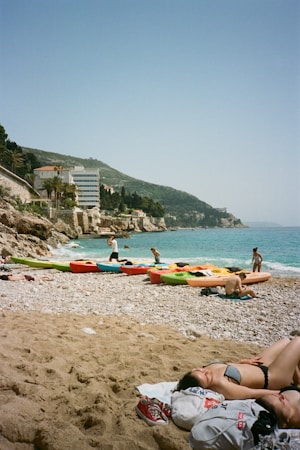 A sunny beach with several kayaks lined up on the shore, people enjoying the water and relaxing on the sand. Hills and buildings provide a scenic backdrop, and the sea is a vibrant blue.