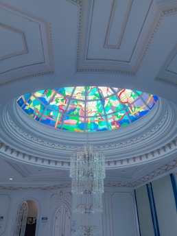 An elegant interior features a large, ornate chandelier suspended from a vibrant stained glass dome ceiling. The ceiling is adorned with intricate patterns in blues, greens, and reds, casting colorful light into the room. The walls and ceiling are detailed with decorative moldings, adding to the opulent atmosphere.