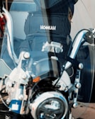 A close-up view of a motorcycle's chrome handlebars and windshield. Behind the windshield, a backpack with the word 'HOOKAH' is visible. The image captures the reflective surfaces and intricate mechanical details of the motorcycle.