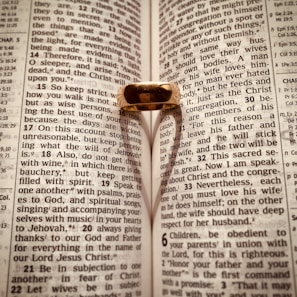 An open Bible with a gold ring placed on it, casting a heart-shaped shadow on the pages. The text is from Ephesians, focusing on themes related to love and wisdom.