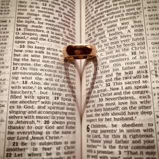 An open Bible with a gold ring placed on it, casting a heart-shaped shadow on the pages. The text is from Ephesians, focusing on themes related to love and wisdom.
