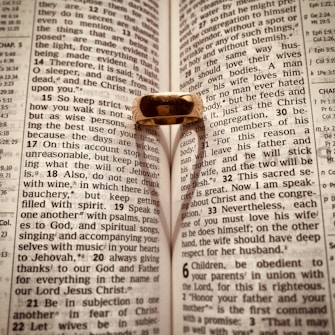 An open Bible with a gold ring placed on it, casting a heart-shaped shadow on the pages. The text is from Ephesians, focusing on themes related to love and wisdom.