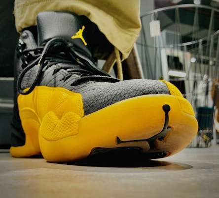 A close-up view of a sneaker with a prominent black and yellow color scheme. The sneaker features textured leather material and has a distinctive logo on the side near the lace area. The shoe is positioned on a smooth surface, and the background includes indistinct elements, possibly indicating an indoor setting.