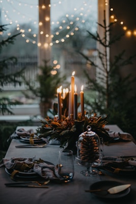 A beautifully set dining table with lit candles at the center, surrounded by elegant tableware and a festive decoration involving greenery and pinecones. The background features large windows adorned with twinkling string lights, adding a cozy, warm ambiance to the setting.