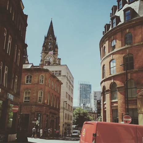 Urban street scene featuring historic architecture, dominated by a tall clock tower with a flag. Red brick buildings frame the street, while modern high-rises are visible in the background. A red van is parked in the foreground, with pedestrians walking nearby.