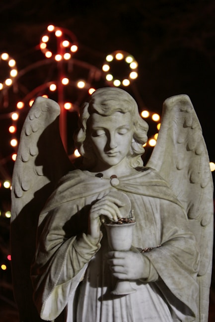 A stone sculpture of an angel holding a chalice, positioned against a dark background. The angel has large wings and a serene expression, with eyes closed. Out of focus, colorful lights form circular patterns behind the statue, enhancing the scene.