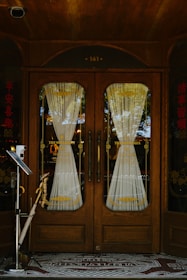 A wooden double door with ornate design and glass panels is adorned with intricate lace curtains. The door is framed by decorative wooden features, complemented by Asian-style text on both sides. A vintage feel is enhanced by the mosaic tile entrance mat that reads 'The Nostalgia'. Reflective surfaces capture bits of the external environment including trees and sky.