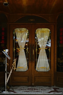 A wooden double door with ornate design and glass panels is adorned with intricate lace curtains. The door is framed by decorative wooden features, complemented by Asian-style text on both sides. A vintage feel is enhanced by the mosaic tile entrance mat that reads 'The Nostalgia'. Reflective surfaces capture bits of the external environment including trees and sky.