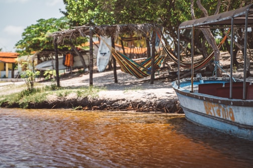 A scenic lakeside view with a small boat docked by the shore and several colorful hammocks hung under a rustic wooden shelter. Surfboards are stacked nearby, and trees provide shade, adding a tranquil atmosphere. A distant building with a red-tiled roof can be seen surrounded by greenery.