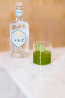 A glass with a green liquid and ice on a marble countertop, next to a bottle labeled Anaë. The background is a warm, neutral tone.