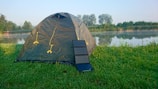 A green dome tent is set up on a grassy area near a calm lake or pond, with trees and bushes in the background. A portable solar panel is positioned against the tent. The early morning or late afternoon light creates a peaceful outdoor setting.