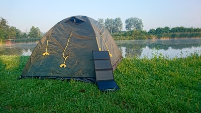 A green dome tent is set up on a grassy area near a calm lake or pond, with trees and bushes in the background. A portable solar panel is positioned against the tent. The early morning or late afternoon light creates a peaceful outdoor setting.