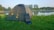 A green dome tent is set up on a grassy area near a calm lake or pond, with trees and bushes in the background. A portable solar panel is positioned against the tent. The early morning or late afternoon light creates a peaceful outdoor setting.