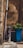 An outdoor scene featuring an old, rustic water pump with intricate designs, situated next to a tall, blue planter containing green foliage. The backdrop is a weathered stone wall with visible texture and signs of age. A large pipe runs vertically along the wall.