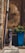 An outdoor scene featuring an old, rustic water pump with intricate designs, situated next to a tall, blue planter containing green foliage. The backdrop is a weathered stone wall with visible texture and signs of age. A large pipe runs vertically along the wall.