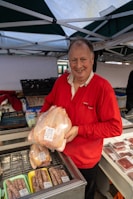 A person in a red shirt is standing behind a market stall displaying various meat products. The individual is holding a packaged whole chicken with price labels visible. The stall contains trays with sausages and other meats, all organized neatly in a display case. Canopies are set up overhead, suggesting an outdoor market setting.