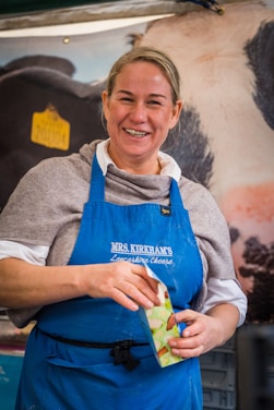 A smiling person wearing a blue apron with the words 'MRS. KIRKHAM'S Lancashire Cheese' written on it, holding a box with a colorful print. The person has light brown hair tied back and is standing in front of a large banner displaying an image of a cow.