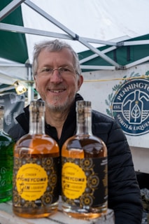 A man with glasses and gray hair stands smiling behind two bottles of gin liqueur. The bottles have decorative labels with a yellow and black design. The backdrop includes a tent and a branding sign that reads 'Pennington Spirits & Liqueur'.
