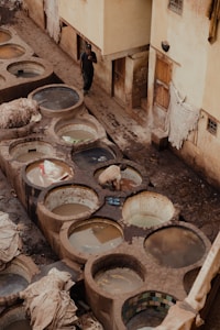 Several large, round stone vats filled with water or dye are located outdoors, surrounded by worn yellowish buildings. A person stands next to a building, while another person kneels by one of the vats, possibly working with the contents. The scene appears to be an outdoor tannery or dyeing facility.