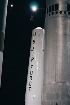 A large rocket labeled 'US Air Force' is positioned in a dark, industrial setting. The rocket is primarily white with bold black lettering, and there are other large structural components visible in the background.
