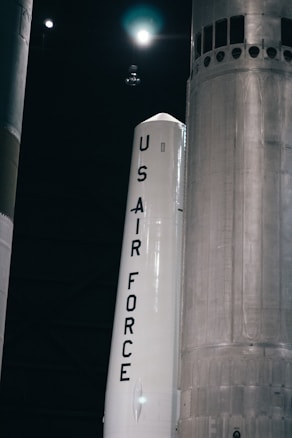 A large rocket labeled 'US Air Force' is positioned in a dark, industrial setting. The rocket is primarily white with bold black lettering, and there are other large structural components visible in the background.