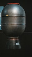 A large metallic object resembling a rocket engine or space propulsion system is displayed against a dark background. It has a cylindrical shape with a slightly rounded top and a flared bottom section. The surface appears smooth and industrial, with several horizontal bands encircling it. There is a small label or sign affixed near the base.