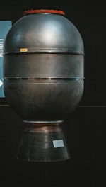 A large metallic object resembling a rocket engine or space propulsion system is displayed against a dark background. It has a cylindrical shape with a slightly rounded top and a flared bottom section. The surface appears smooth and industrial, with several horizontal bands encircling it. There is a small label or sign affixed near the base.