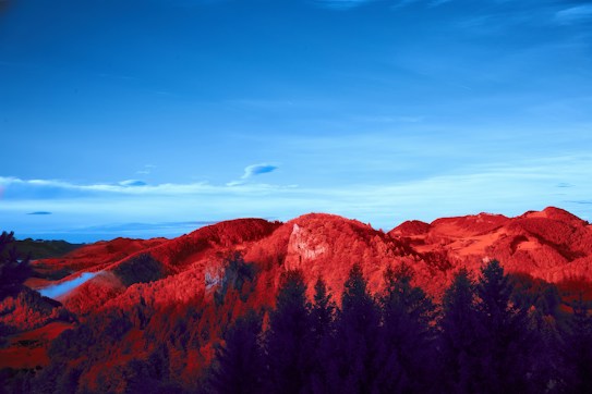 A surreal landscape featuring red-hued mountains and dark purple trees under a vibrant blue sky. Clouds appear in the sky, enhancing the otherworldly atmosphere.