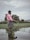 A person stands in shallow water in a flooded agricultural field, wearing a pink shirt and black shorts. In the background, there is a large, partially obscured building, possibly a house, surrounded by greenery. The sky is overcast, adding a grayish tone to the scene.
