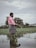 A person stands in shallow water in a flooded agricultural field, wearing a pink shirt and black shorts. In the background, there is a large, partially obscured building, possibly a house, surrounded by greenery. The sky is overcast, adding a grayish tone to the scene.
