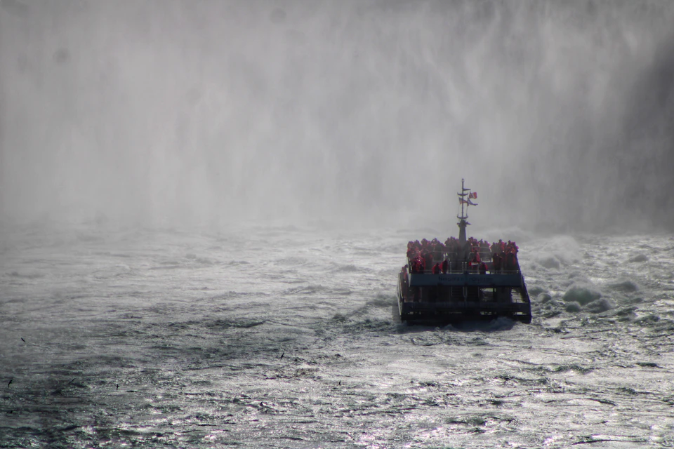 The image depicts a boat sailing on a river, with a waterfall cascading down behind it. The boat is filled with passengers and appears to be moving towards the right side of the frame. The water surrounding the boat has a grayish hue, suggesting that it might be a cloudy or overcast day.