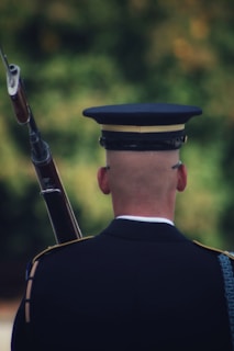 A person in a formal military uniform is seen from behind, holding a rifle. The person wears a dark uniform with gold accents and a hat. The background is blurred with green and hints of orange, suggesting an outdoor setting.