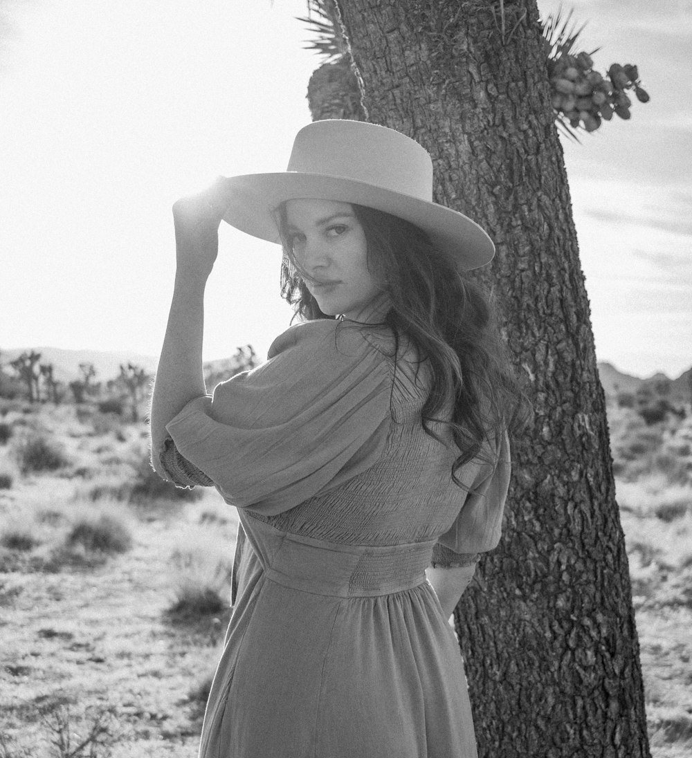 a woman wearing a hat and standing next to a tree