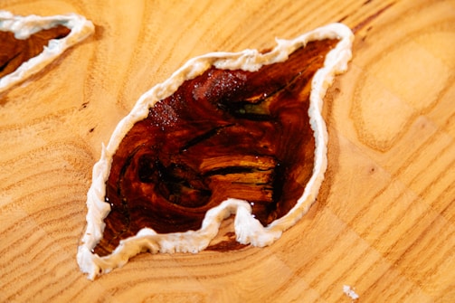 A wooden surface features an abstract design with a glossy, brown, leaf-shaped inlay bordered by a creamy white edge. The wood grain is visible and textured, adding a natural and artistic element to the composition.