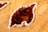 A wooden surface features an abstract design with a glossy, brown, leaf-shaped inlay bordered by a creamy white edge. The wood grain is visible and textured, adding a natural and artistic element to the composition.