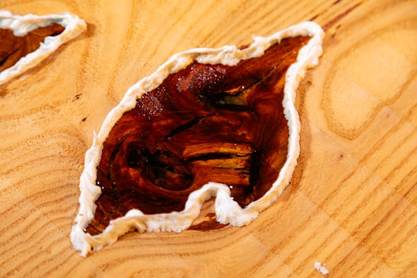 A wooden surface features an abstract design with a glossy, brown, leaf-shaped inlay bordered by a creamy white edge. The wood grain is visible and textured, adding a natural and artistic element to the composition.