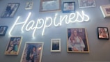 A collection of photographs is arranged on a wall, centered around a glowing neon sign that spells out the word 'Happiness'. The photographs feature various individuals, suggesting a personal or family display.
