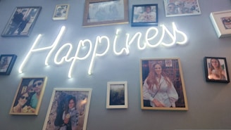A collection of photographs is arranged on a wall, centered around a glowing neon sign that spells out the word 'Happiness'. The photographs feature various individuals, suggesting a personal or family display.