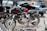 A vintage Yamaha motorcycle with a Vindicator front fairing is parked amidst several cars with open trunks in a scrapyard. The motorcycle appears aged and possibly inoperative, surrounded by spare parts and debris. The scene suggests an atmosphere of decay and abandonment, with various car parts strewn across the gravel ground.