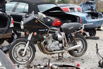 A vintage Yamaha motorcycle with a Vindicator front fairing is parked amidst several cars with open trunks in a scrapyard. The motorcycle appears aged and possibly inoperative, surrounded by spare parts and debris. The scene suggests an atmosphere of decay and abandonment, with various car parts strewn across the gravel ground.