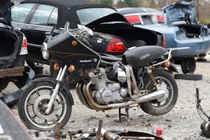 A vintage Yamaha motorcycle with a Vindicator front fairing is parked amidst several cars with open trunks in a scrapyard. The motorcycle appears aged and possibly inoperative, surrounded by spare parts and debris. The scene suggests an atmosphere of decay and abandonment, with various car parts strewn across the gravel ground.