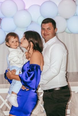A family of three is posing in front of a background decorated with white and light blue balloons. The mother, dressed in a blue off-shoulder dress, is holding and kissing their baby, who is dressed in a white shirt and light blue pants. The father, wearing a white shirt and dark pants, is standing close to them, smiling at the camera.