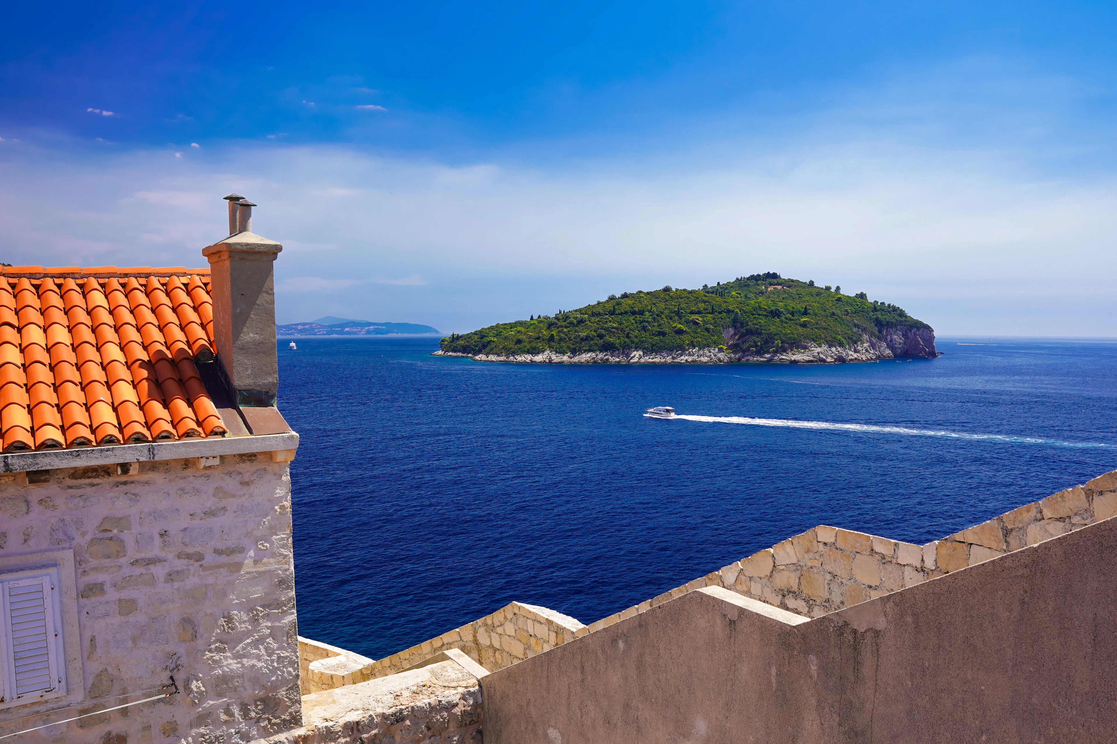 A terracotta rooftop and chimney frame a view of Lokrum island and a passing boat off Dubrovnik.
